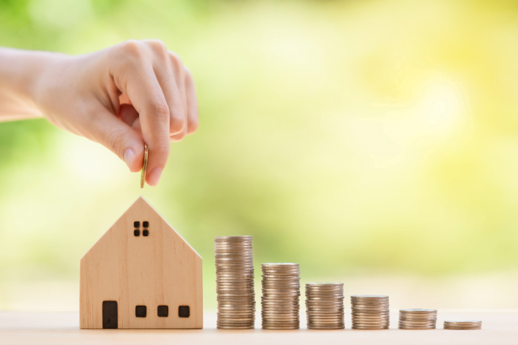 A hand is putting coins into a wooden house model, signifying an increase in property value.