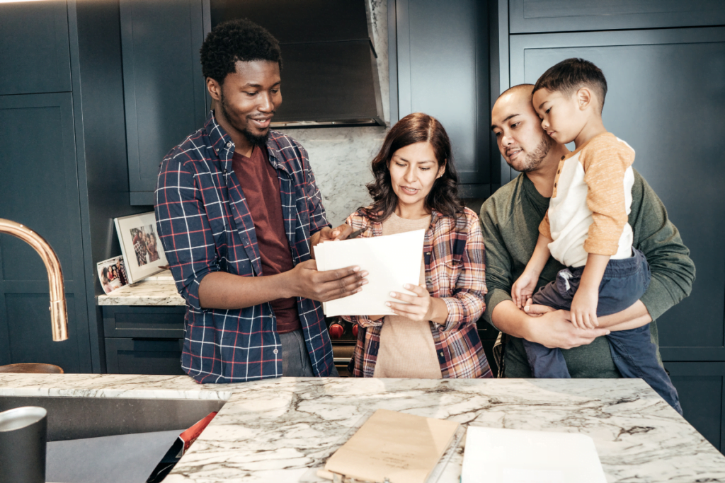 A group of four people, including a child being held, are gathered in a modern kitchen looking at a document.