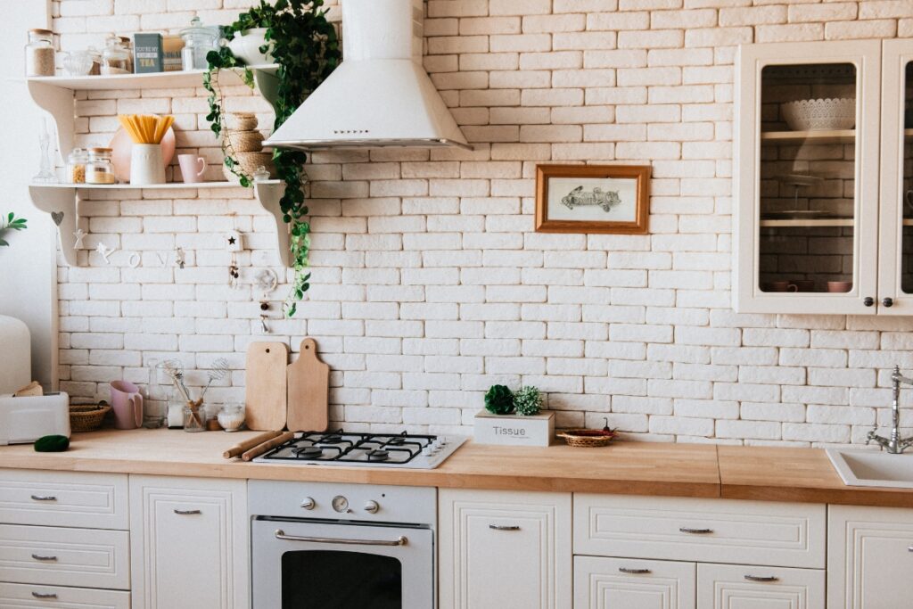 A cozy kitchen interior featuring white cabinetry, a brick backsplash, wooden countertops, and a stove with hanging green plants offers practical kitchen maintenance tips.