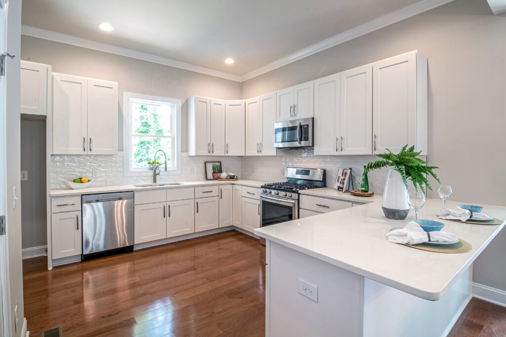 Modern kitchen maintenance tips suggest white cabinetry, stainless steel appliances, and an island with dining setup should be illuminated by natural light from a window for enhanced aesthetics and energy efficiency.