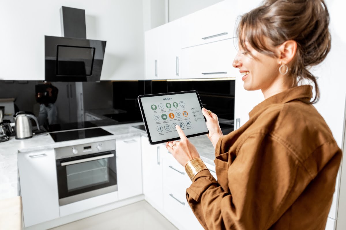 A woman in a brown jacket uses a tablet with a smart kitchen technology interface in a modern kitchen.