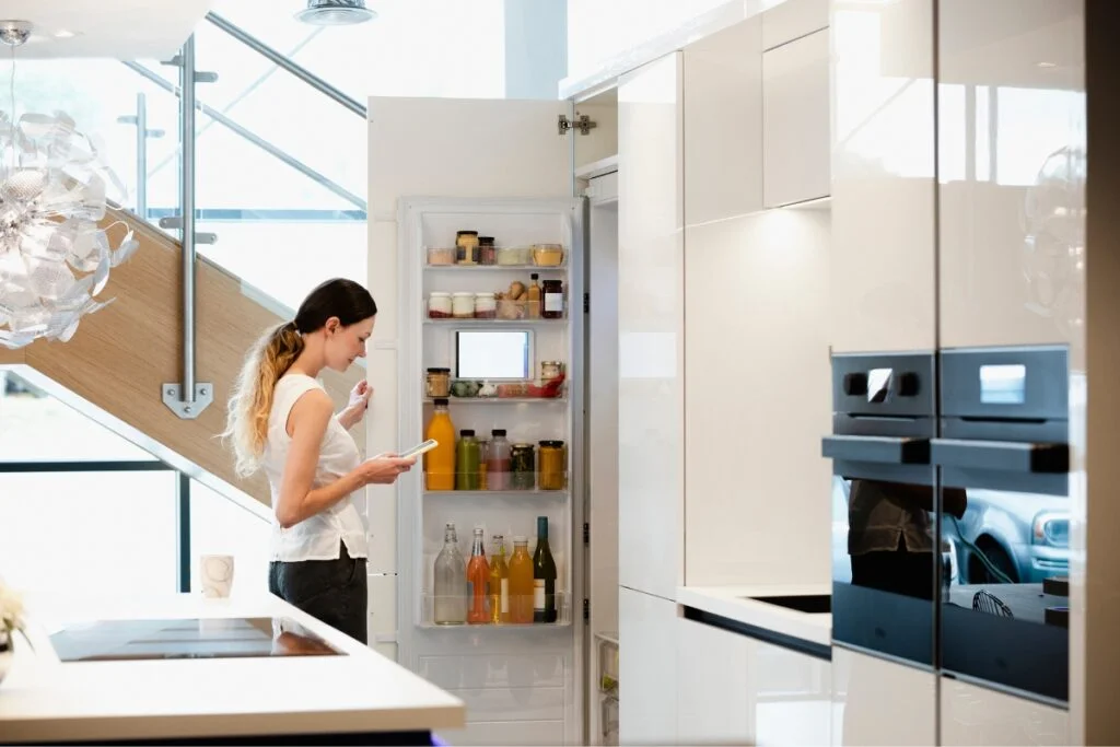 Smart Kitchen Technology: Revolutionizing Kitchens with Home Tech Integration 2 A woman in a smart kitchen examines the contents of an open refrigerator while holding a smartphone.