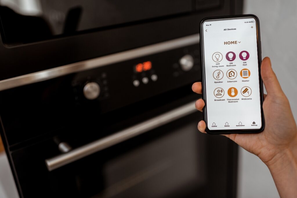 A person's hand holding a smartphone with a smart kitchen technology app open, in front of a modern oven.