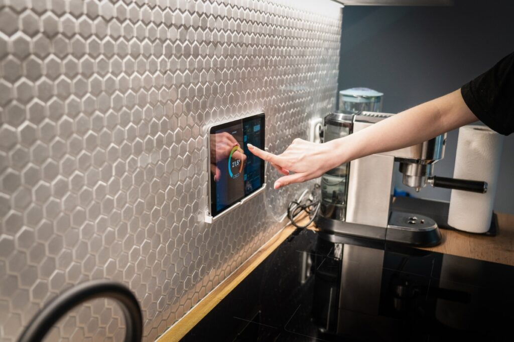 A person's hand adjusting a wall-mounted digital tablet displaying a timer in a smart kitchen with hexagonal tiles.