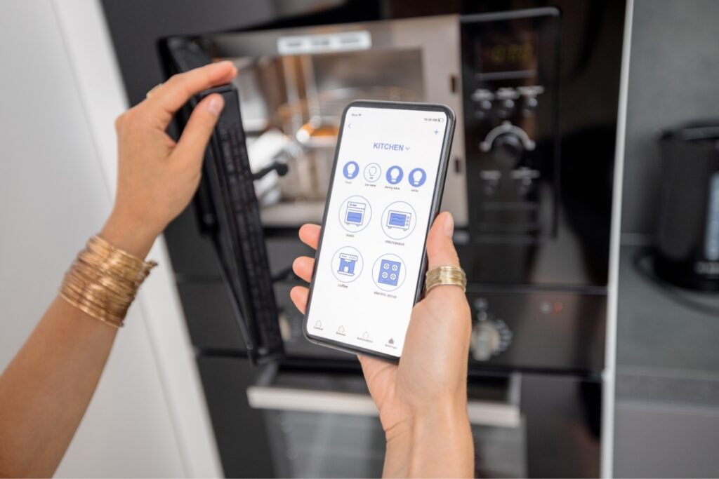 A person uses a Smart Kitchen Technology app to control kitchen appliances, with a microwave and oven in the background.