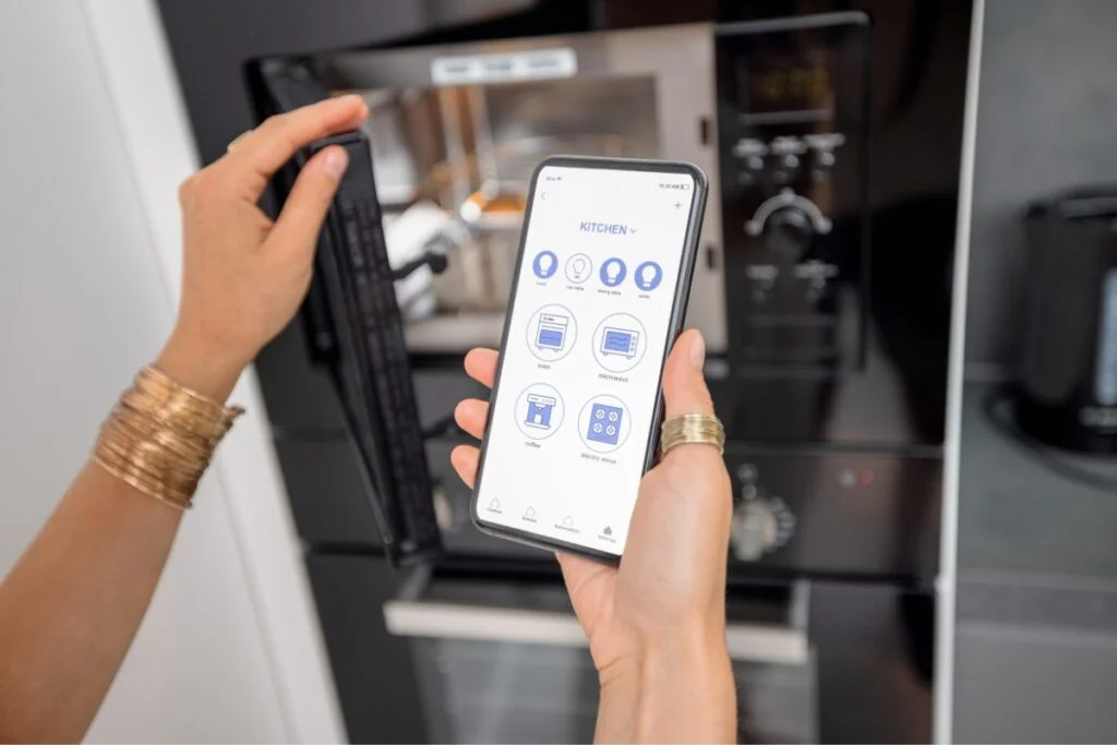 Smart Kitchen Technology: Revolutionizing Kitchens with Home Tech Integration 6 A person uses a Smart Kitchen Technology app to control kitchen appliances, with a microwave and oven in the background.