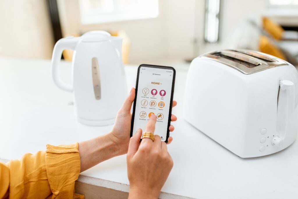 A person uses a smartphone to control Smart Kitchen Technology, including a kettle and a toaster, through a home automation app.