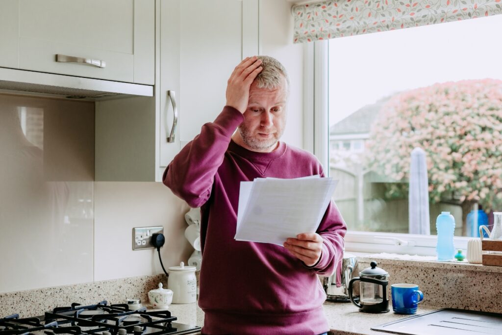 A man stands in a kitchen looking puzzled while holding a piece of paper about home energy efficiency. He has one hand on his head and the other holding the paper. The kitchen has light cabinets and a window with a floral blind.