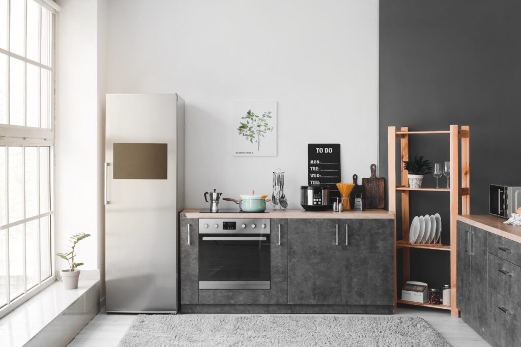 A modern kitchen with a stainless steel refrigerator, stove, and grey cabinets. A light teal pot sits on the stove. Shelves hold plates and glasses. Natural light enters through large windows, enhancing the home energy efficiency of this stylish space.