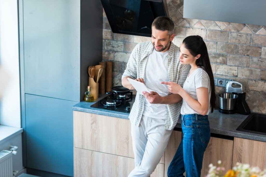 A man and woman stand in a kitchen looking at a tablet. The woman points at the screen while they discuss something, possibly about home energy efficiency. The kitchen has modern decor with wooden cabinets and a tiled backsplash.