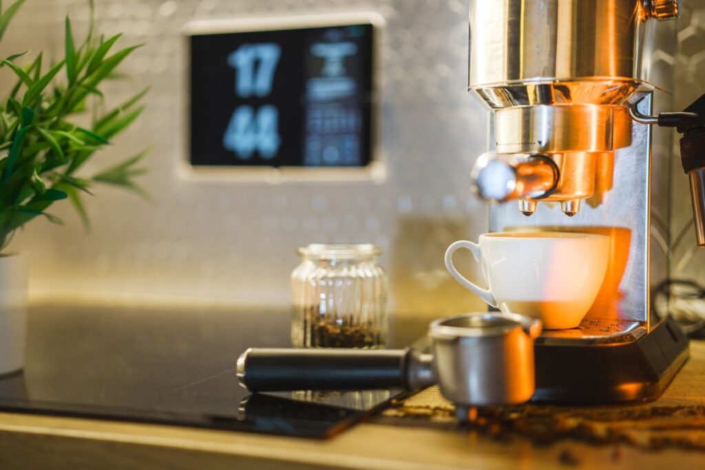 A modern espresso machine brewing coffee into a white cup, with a portafilter and a jar of coffee beans nearby, embodies contemporary kitchen design trends. A digital clock displaying 17:44 is in the background, while a plant adds a touch of greenery on the left.