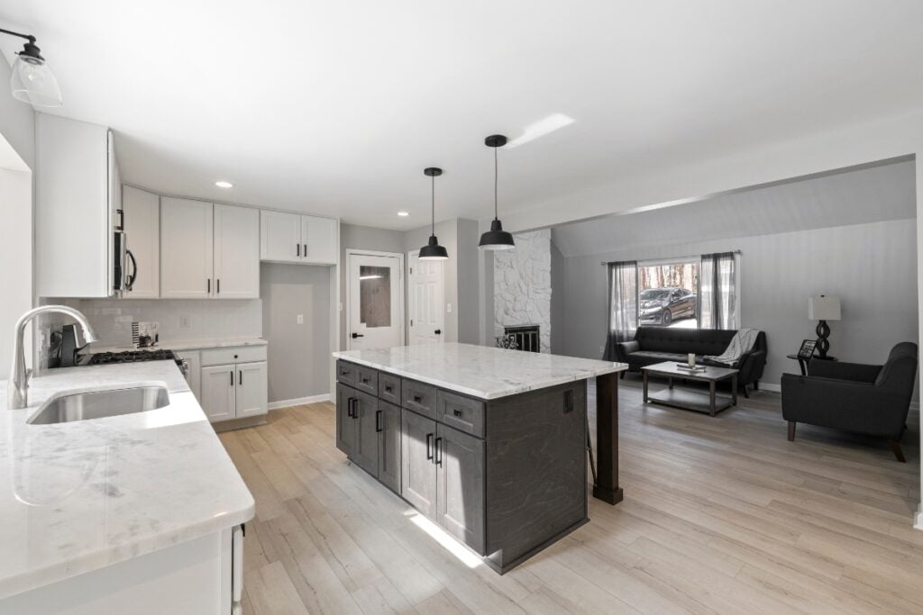 Modern kitchen with white cabinets, a large center island with a marble countertop, pendant lights, stainless steel appliances, and an open view into a living area with a couch and a lamp—a perfect embodiment of current kitchen design trends.