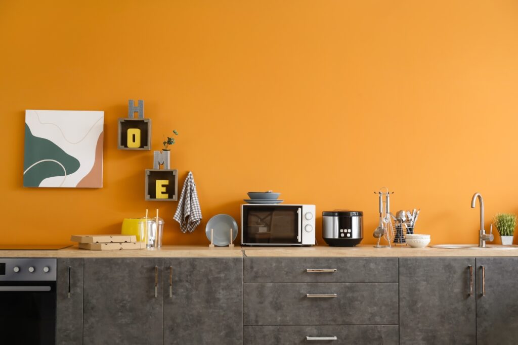 Modern kitchen with concrete countertops, an orange wall, and various appliances including a microwave, coffee maker, and kettle. Embracing the latest kitchen design trends, decorative "HOME" letters and artwork hang above the counter.