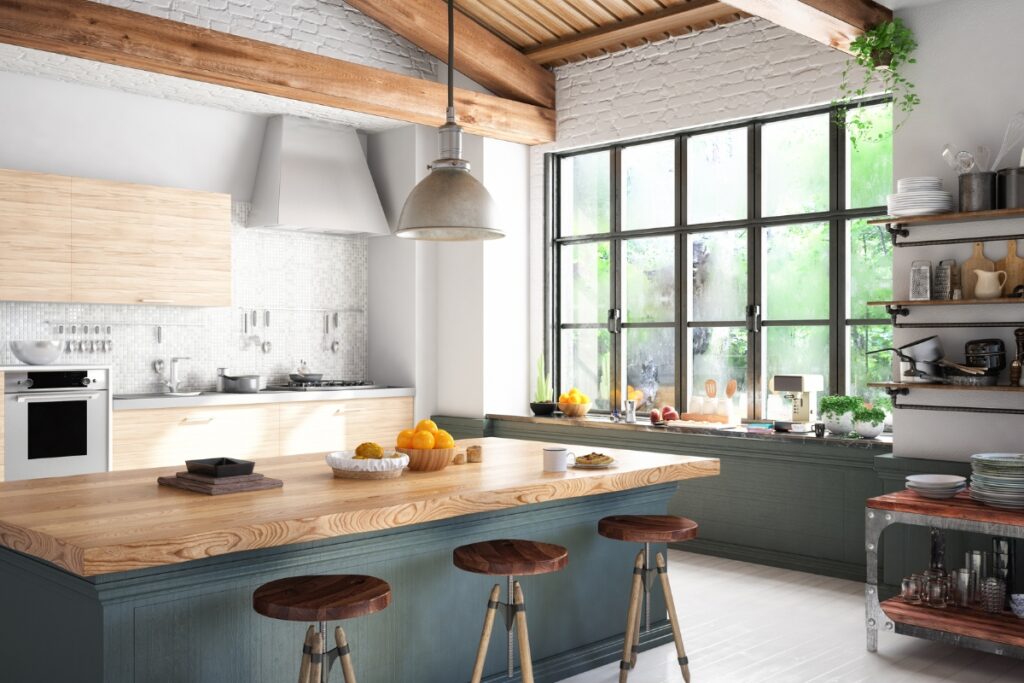 Modern kitchen with large windows, a wooden island with three stools, and various kitchen items. White walls, wooden beams, and greenery visible outside the window reflect the latest kitchen design trends.