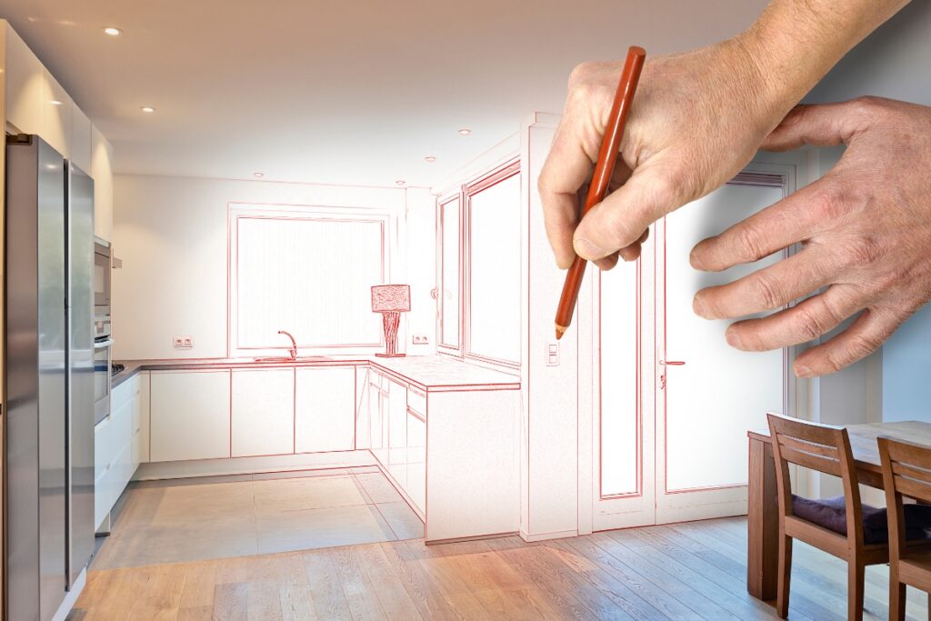 A hand holding a pencil is drawing a kitchen in red lines overlaid on a photograph of a modern Atlanta kitchen with wooden floors, white cabinets, and a dining table with chairs, illustrating an innovative real estate renovation.