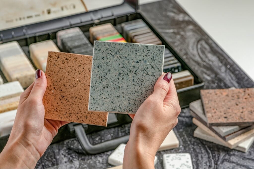 Two hands hold up mosaic tile samples, one brown and one gray, in front of an open sample case filled with various other tiles. This selection plays a crucial role in the Atlanta real estate renovation scene, adding both style and value to homes.