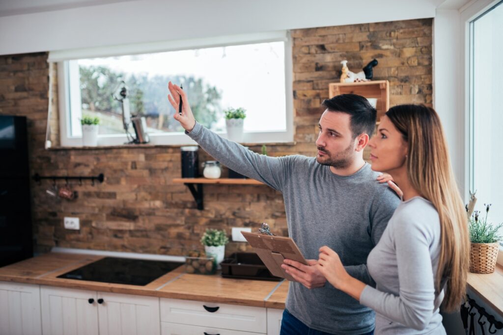 A man and a woman stand in a kitchen, with the man gesturing towards something out of frame. The woman looks in the same direction, intrigued. The man holds a clipboard, likely discussing their next steps in an Atlanta real estate renovation project.