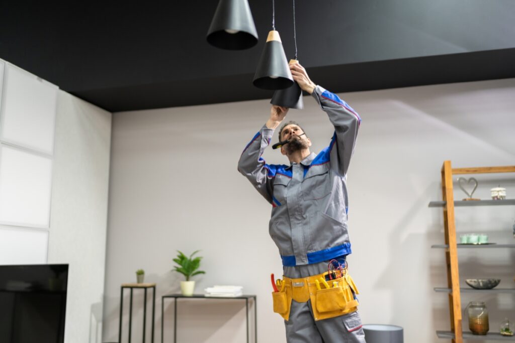 A worker in a gray uniform with a yellow tool belt is standing on a ladder, fixing or installing black pendant lamps as part of an Atlanta real estate renovation in a modern interior setting.