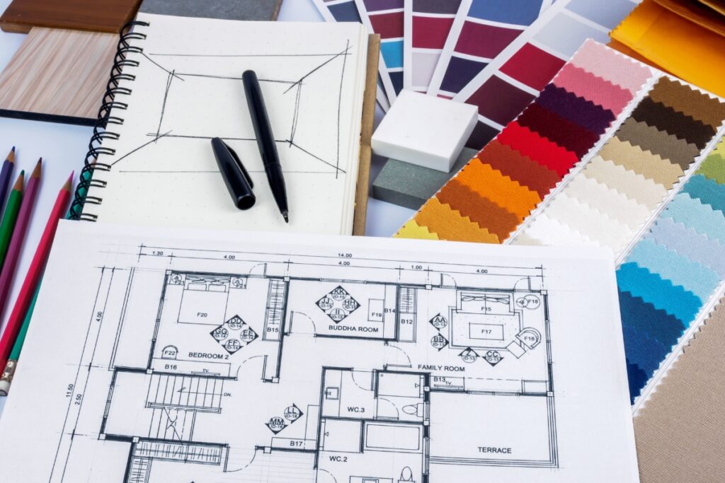 Architectural floor plans, a notebook with a pen, color swatches, fabric samples, and colored pencils arranged on a table suggest an Atlanta real estate renovation taking shape through meticulous interior design planning.