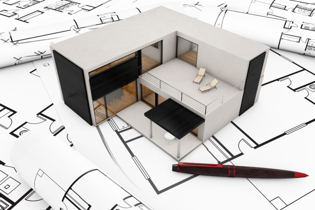 A 3D model of a modern house with balconies and large windows, placed on top of several architectural blueprints. A red and black pen is positioned in the foreground, hinting at an Atlanta real estate renovation project.