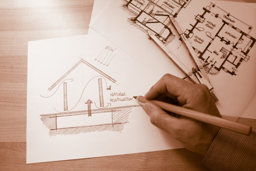 A hand draws a sketch of a house's natural ventilation system on paper, with architectural plans and a compass placed on the desk, showcasing an ambitious Atlanta real estate renovation project.