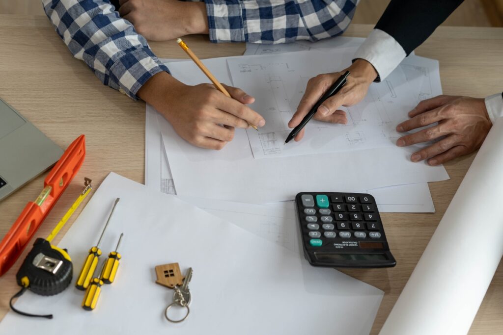 Two people reviewing architectural plans on a table with a calculator, pencils, a tape measure, screwdrivers, keys, and other drafting tools nearby as they discuss an Atlanta real estate renovation.