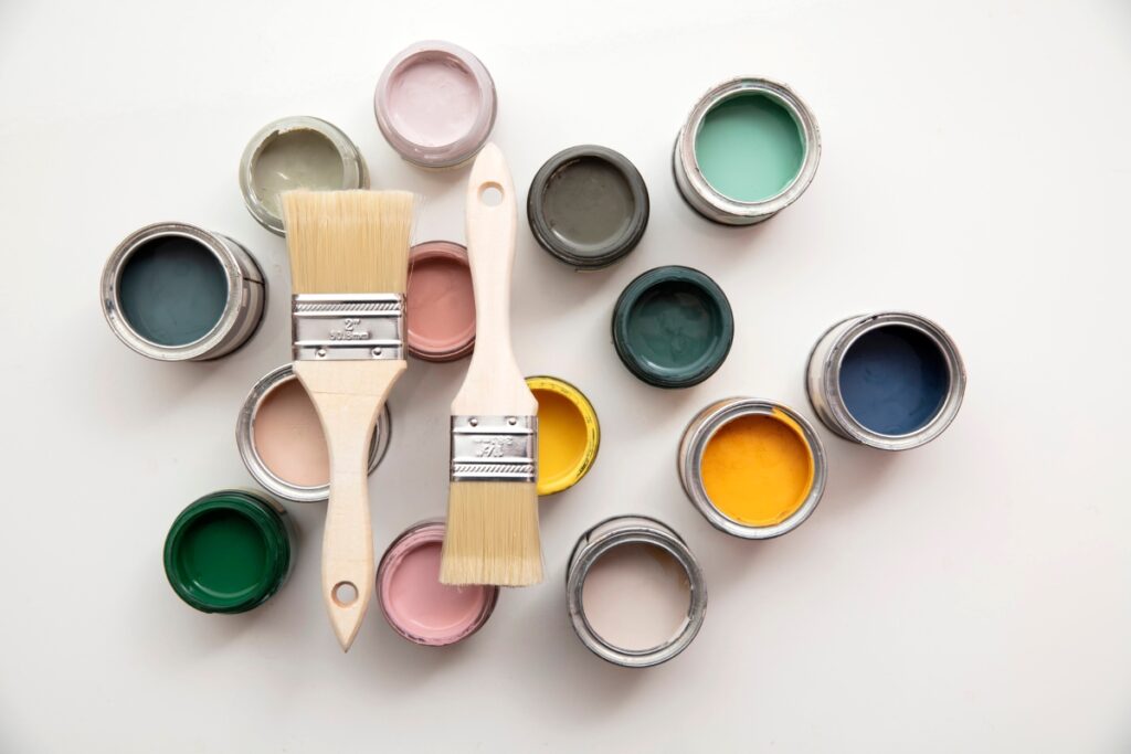 Top view of open paint cans of various colors and two paintbrushes arranged on a white surface, perfect for inspiring your DIY bathroom projects.