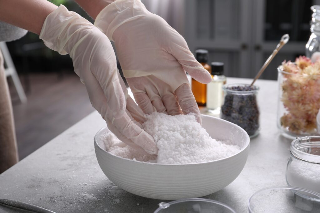 Hands in gloves holding white powder in a bowl, surrounded by jars and bottles on a table, perfect for diy bathroom projects.