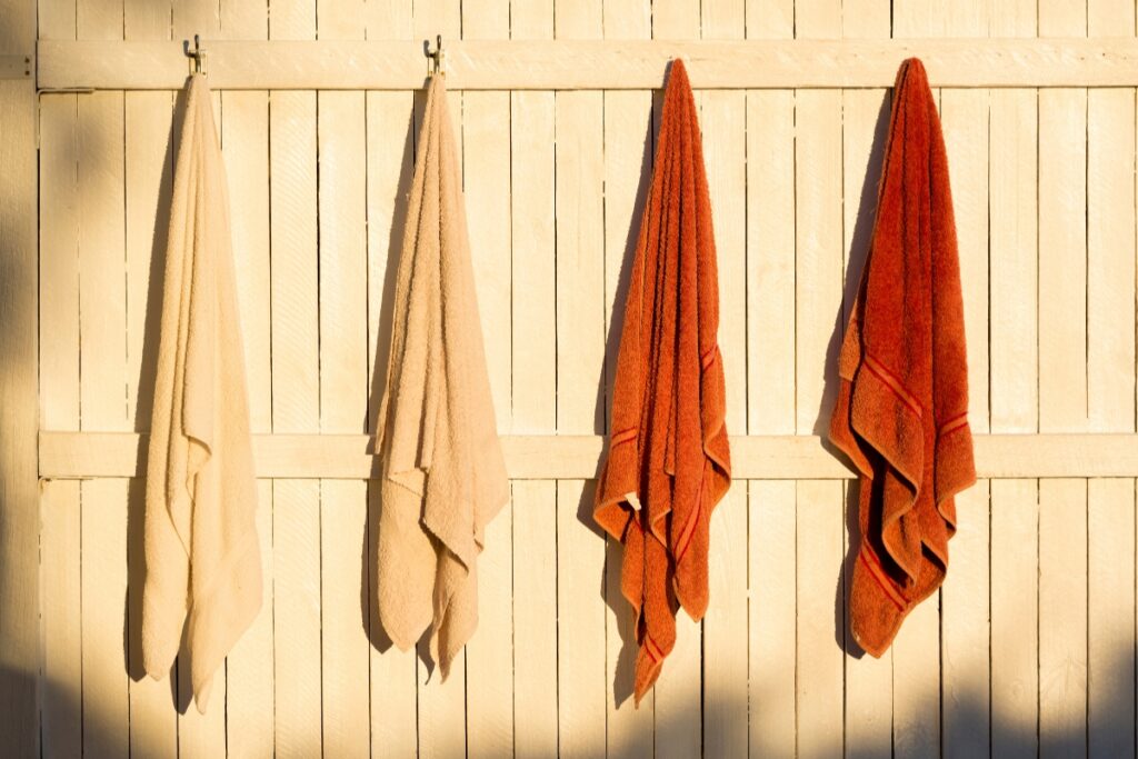 Four towels, two beige and two rust-colored, hang on hooks attached to a light wooden fence in the sunlight, showcasing a simple yet effective idea for DIY bathroom projects.