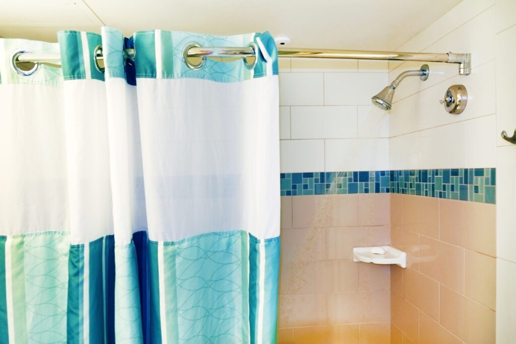 A white-tiled shower with a blue and white shower curtain. The shower, a perfect subject for diy bathroom projects, features a single showerhead and a small shelf holding soap.