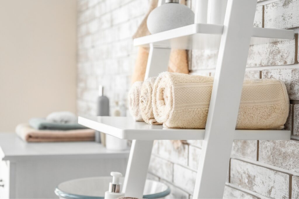 Two rolled beige towels on a white ladder shelf against a white brick wall, with additional toiletries and towels blurred in the background, create an inviting touch for your DIY bathroom projects.