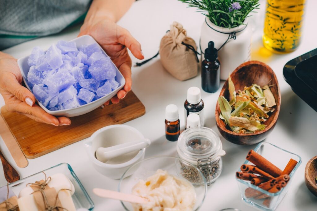 A person holding a bowl of blue soap cubes surrounded by various natural ingredients and essential oils on a table, perfect for DIY bathroom projects.