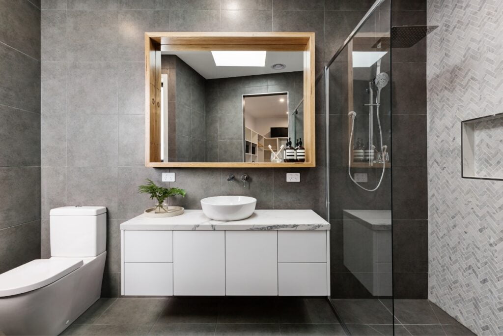 Modern bathroom featuring a rectangular mirror, vessel sink on a white vanity, grey tiles, a toilet, and a glass-enclosed shower area with a rain shower head. Perfect for those inspired by DIY bathroom projects.