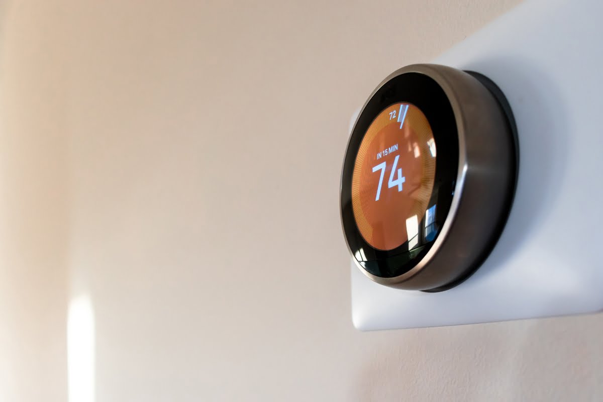 A smart thermostat, integrated into home automation systems, is mounted on a white wall and displays a temperature setting of 74 degrees Fahrenheit.