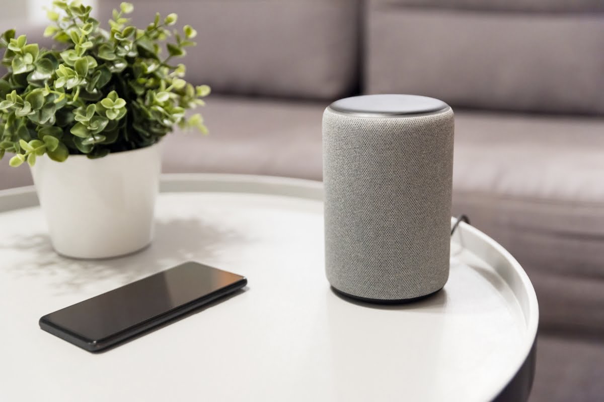 A smart speaker, smartphone, and potted plant on a white round table with a gray couch in the background showcase the elegance of modern home automation systems.