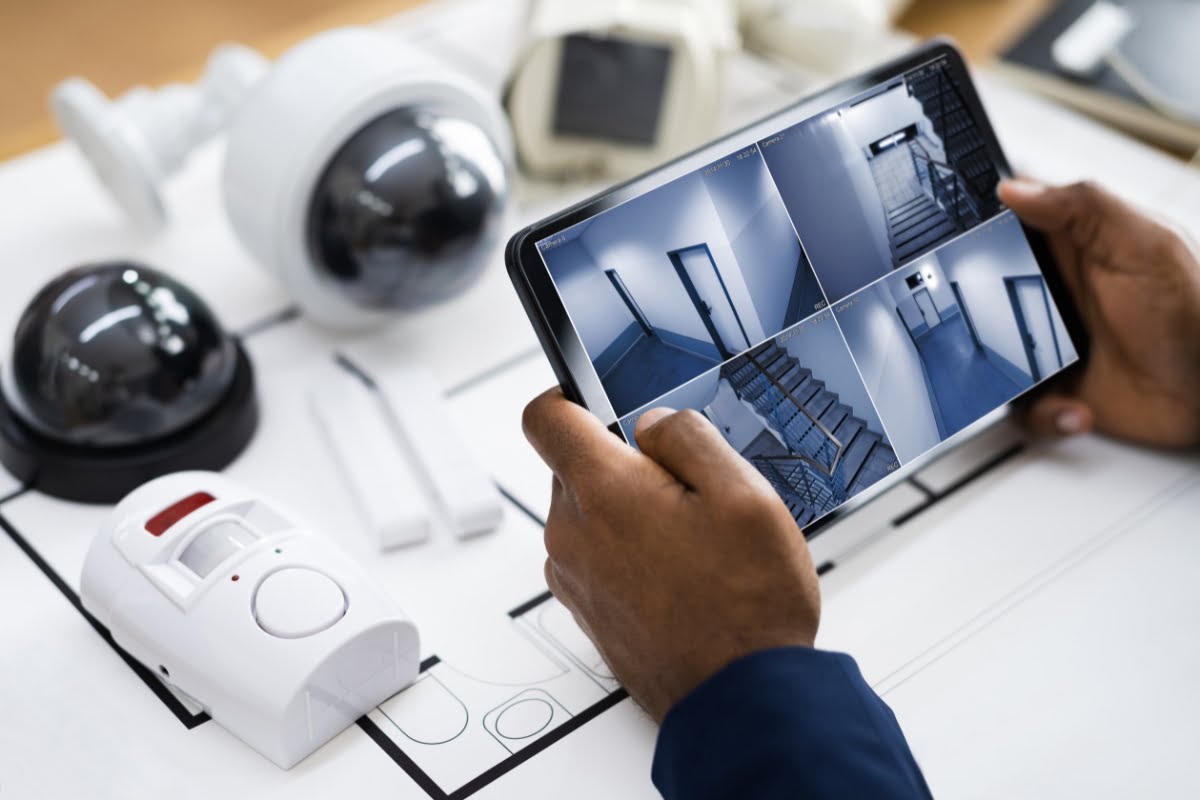 A person views surveillance camera feeds on a tablet, with various security equipment, including cameras and a motion sensor, placed on a desk below, seamlessly integrated into advanced home automation systems.