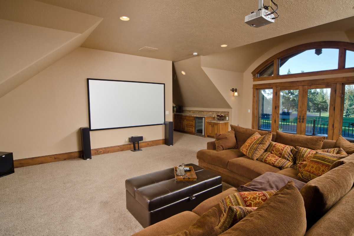 A well-lit home theater room with a large screen, brown sectional sofa, ottoman, and wall-mounted projector. The space features home automation systems for seamless control of lighting and sound. There are large windows and a bar counter in the background.