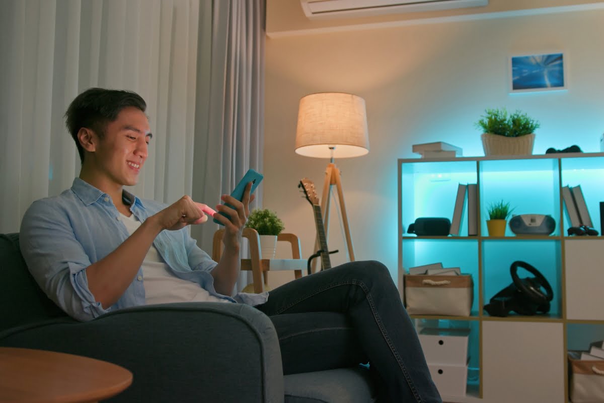 A man sits on a couch in a modern living room, smiling while using his smartphone to control home automation systems. A bookshelf with various items and a lit floor lamp are in the background.