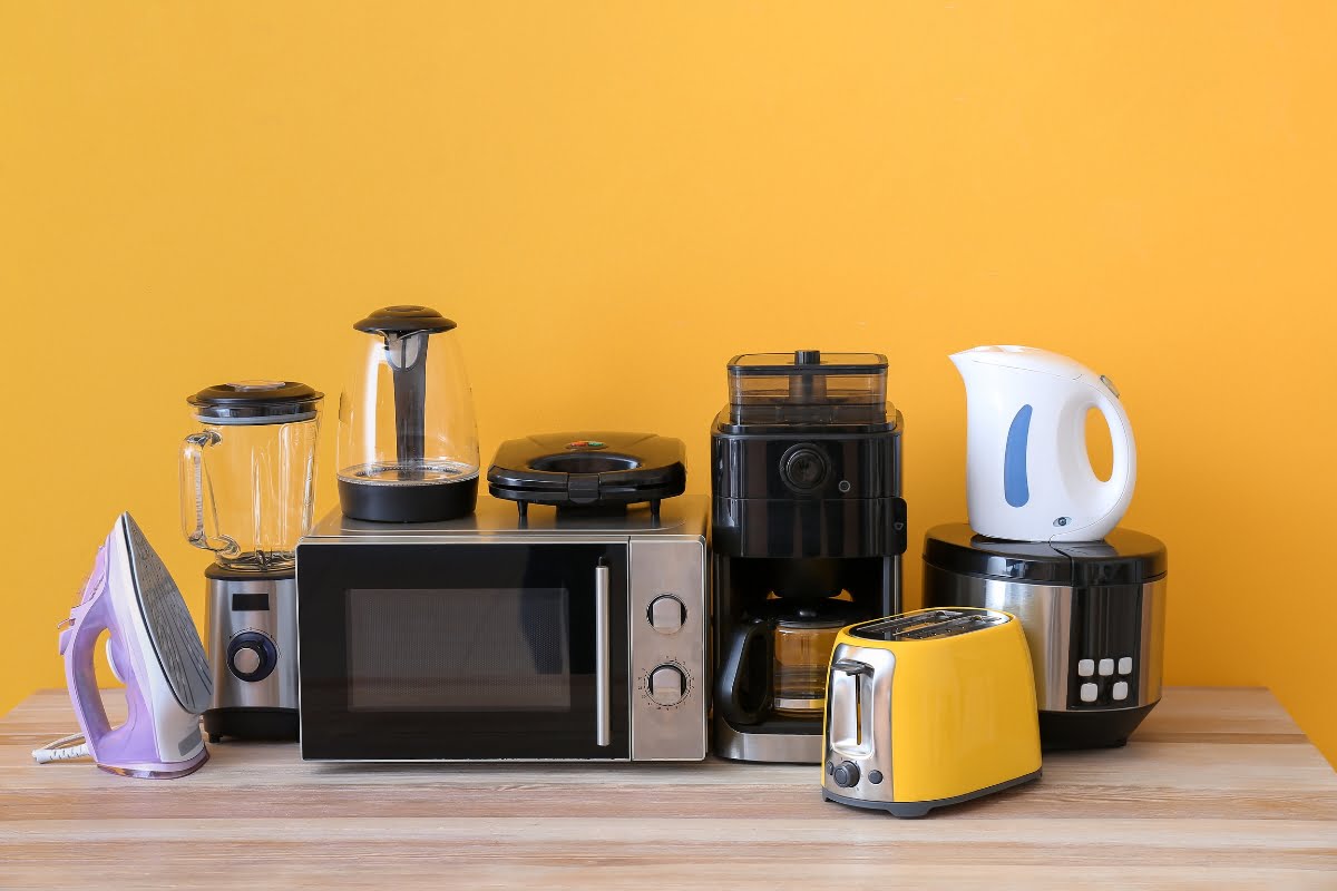 A variety of kitchen appliances, including a blender, iron, microwave, electric kettle, coffee maker, rice cooker, toaster, and food processor, are arranged on a wooden surface against a yellow wall. These essentials can be seamlessly integrated into modern home automation systems for added convenience.