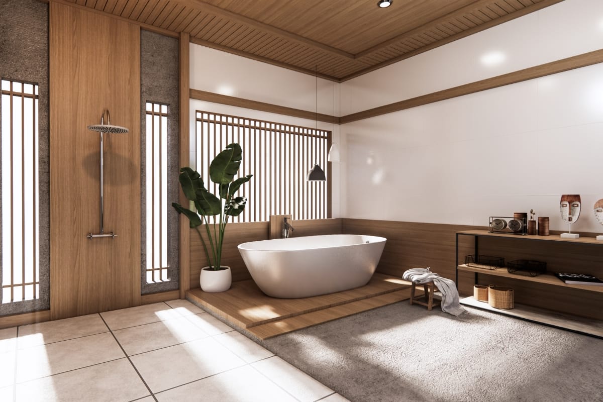 A modern bathroom with a freestanding bathtub, a large plant, wooden accents, and a walk-in shower features cultural design elements. Various accessories on a shelf add character while natural light streams in through vertical wooden slats.