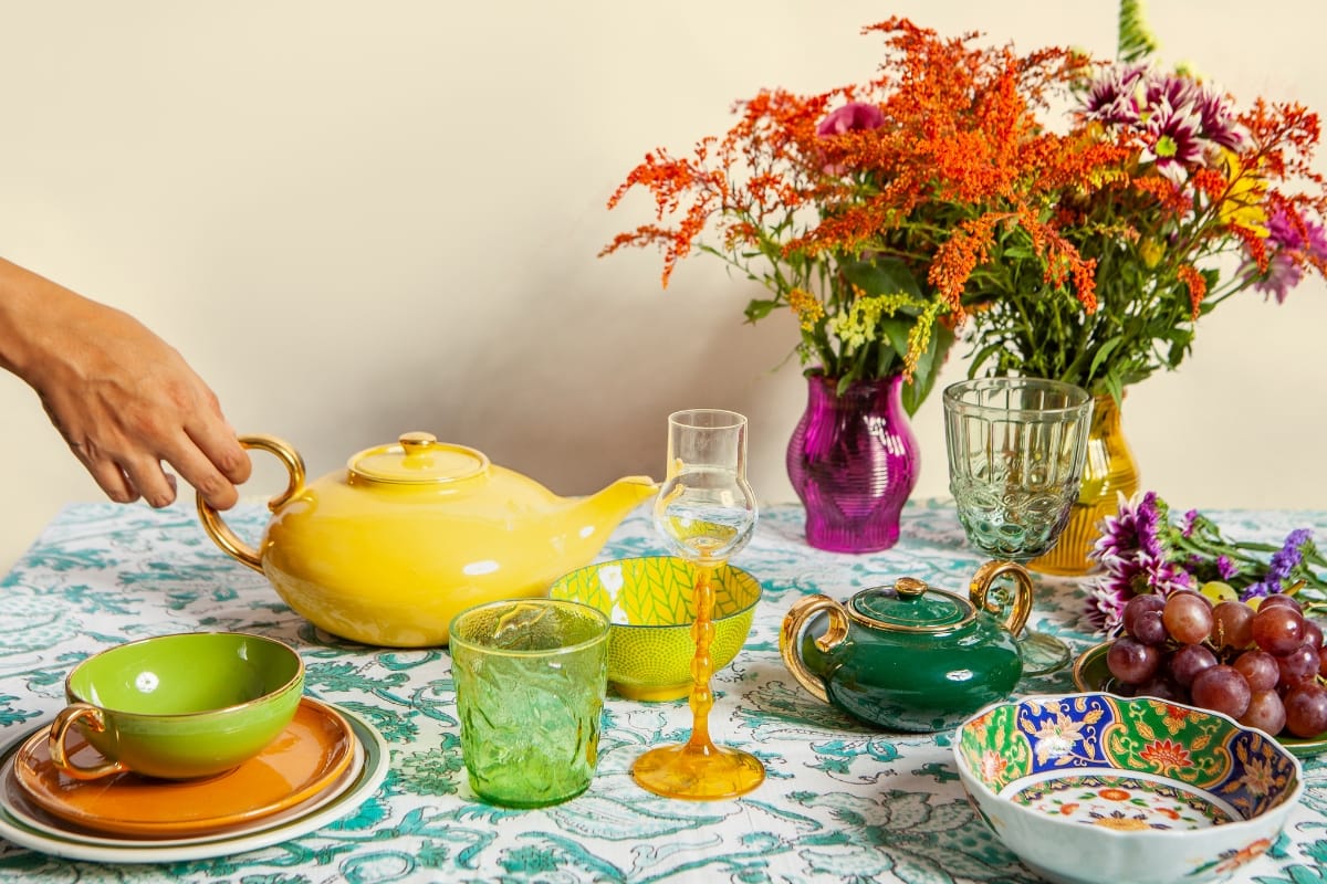 A vibrant table setting with a variety of colorful dishes, glassware, a yellow teapot, green teacups, fresh flowers in vases, and a bunch of grapes. A hand is holding the yellow teapot's handle, blending elements of cultural design throughout.