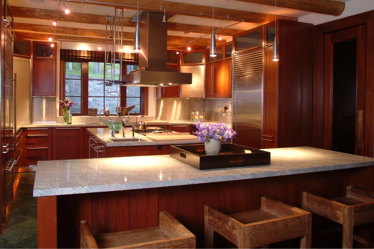 A modern kitchen with wooden cabinetry, a central island boasting a granite countertop, and wooden bar stools. Hanging pendant lights and stainless steel appliances enhance the space, while subtle cultural design touches add character. A vase with purple flowers graces the island.