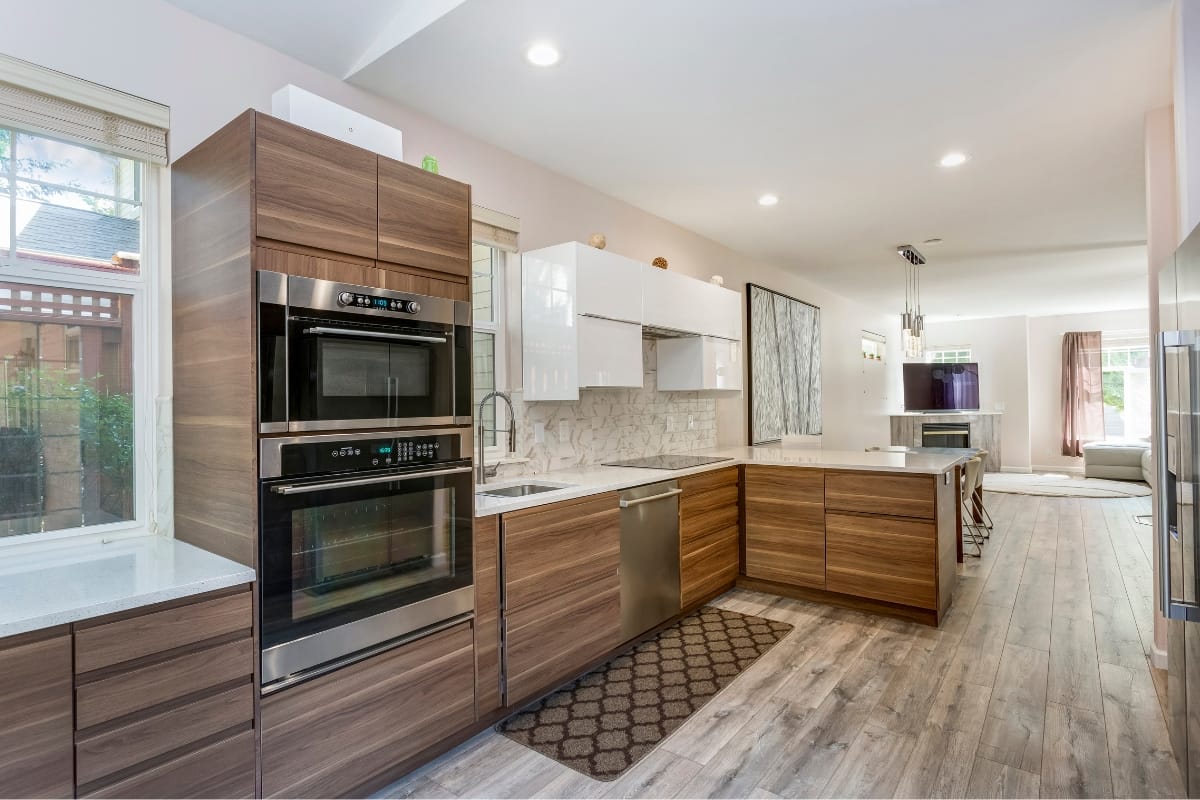 A modern kitchen with wood cabinets, stainless steel double ovens, a dishwasher, and a marble-tiled backsplash. Featuring cultural design elements, there is a center island and an adjoining open dining and living area.