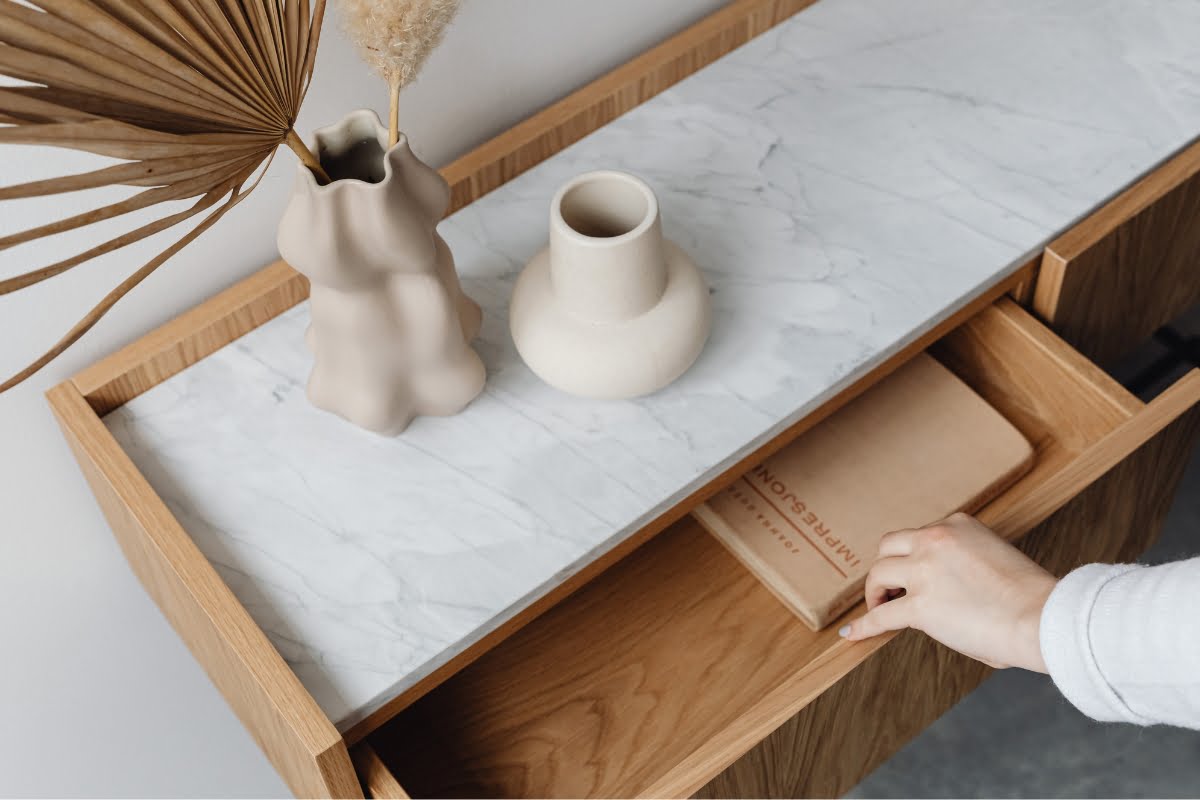 A hand reaches to open a wooden drawer below a marble-top counter, which holds two decorative vases and a book titled "Human Emotion" – a perfect touch for those who appreciate healthy home design.