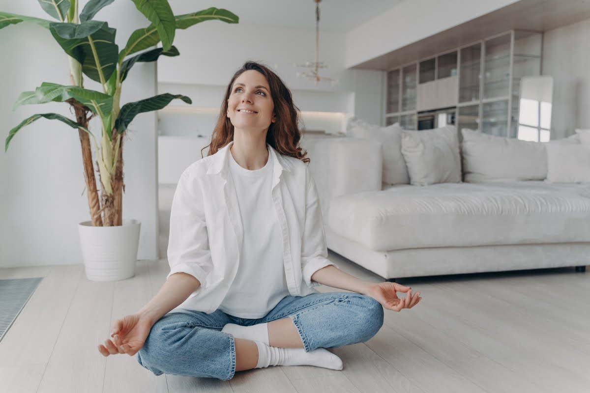 A woman sits cross-legged on the floor in a bright, modern living room, performing a meditation pose with a serene expression. Emphasizing healthy home design, a large potted plant and a white couch create an inviting backdrop.