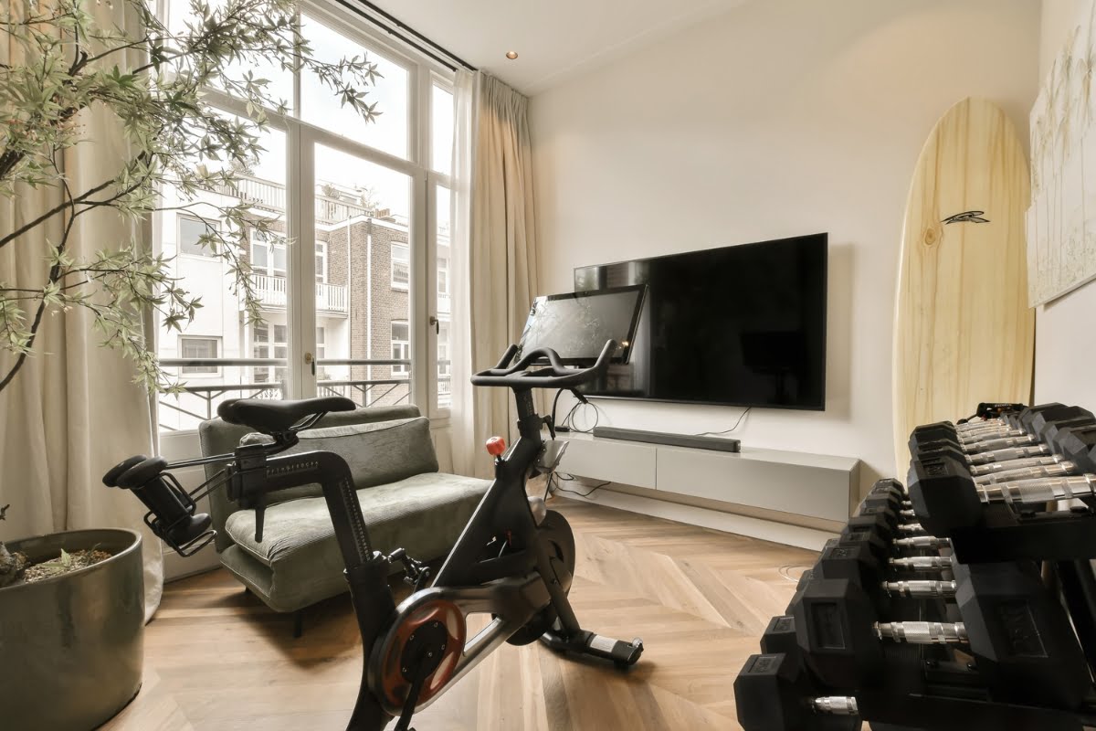 A modern indoor fitness area featuring an exercise bike, dumbbells, a surfboard, and a wall-mounted TV near large windows overlooking a cityscape embraces healthy home design.