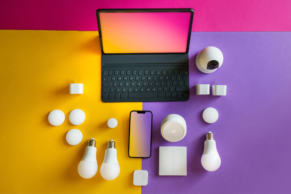 Colorful overhead view of various smart home devices, including a laptop, smartphone, smart bulbs, sensors, and cameras arranged on a yellow and purple background, showcasing the essence of healthy home design.