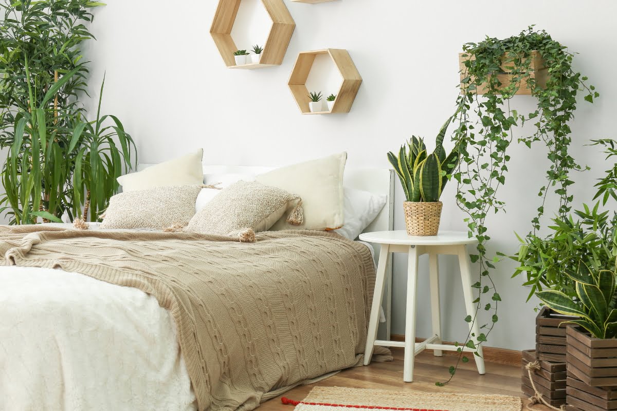 A cozy bedroom features a neatly made bed with beige and white linens, surrounded by various potted plants and hexagonal wooden wall shelves. Embracing healthy home design, a small white side table holds a potted snake plant.