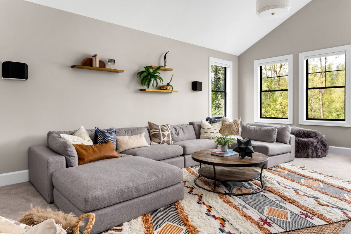 Spacious living room with a large gray sectional sofa, patterned rug, and round coffee table. Wall shelves hold decorations, promoting a healthy home design. Large windows provide ample natural light.