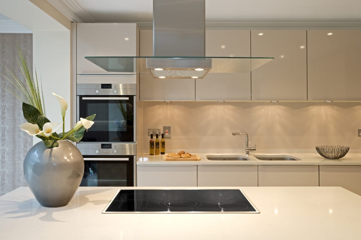 Modern kitchen featuring beige cabinets, dual wall ovens, and an electric cooktop on an island, complemented by a stainless steel range hood and sleek kitchen appliances. Decor includes a large vase with white flowers and a bowl of fruit, adding elegance to the space.